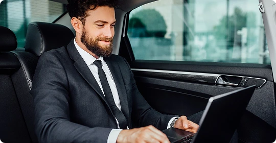 Smiling businessman working on a laptop in the backseat of a luxury sedan, highlighting productivity during chauffeured travel.