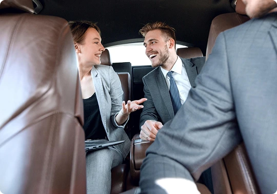 Male and female professionals smiling and conversing inside the back of a luxury car, representing enjoyable and executive group travel.