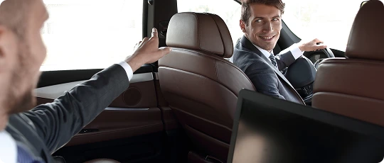Driver smiling at a business passenger while seated inside a luxury vehicle, emphasizing comfort and high-end business transport.